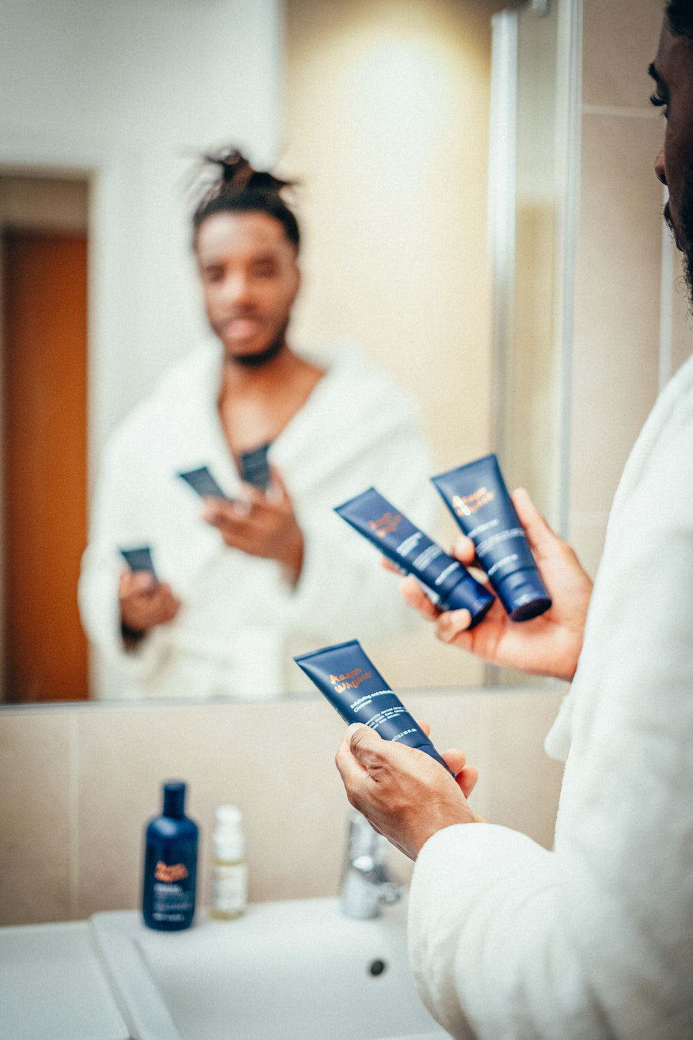 Person in a bathroom holding skincare products in front of a mirror